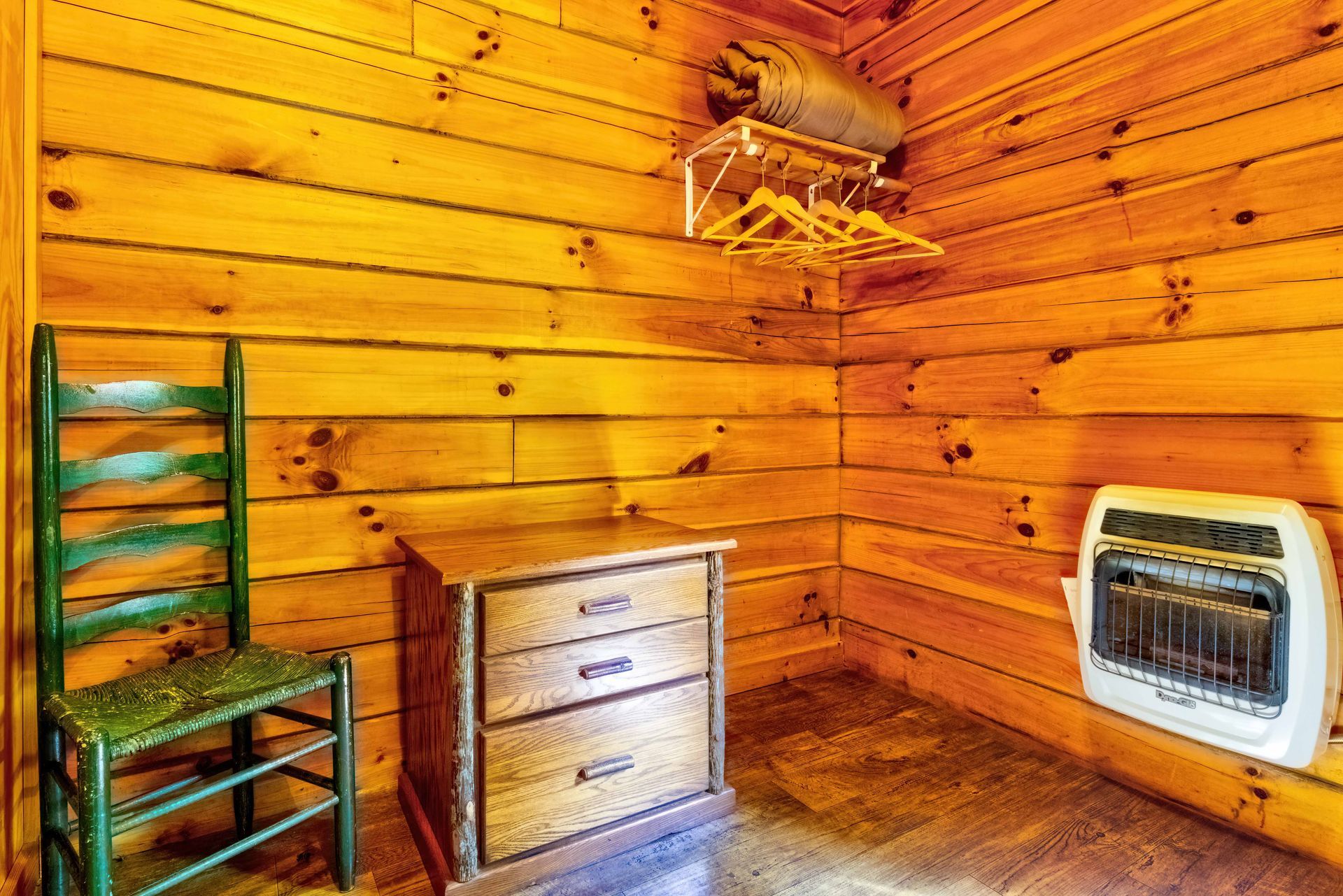 Wooden cabin corner with a green chair, small dresser, wall-mounted rack, and heater by the wall.