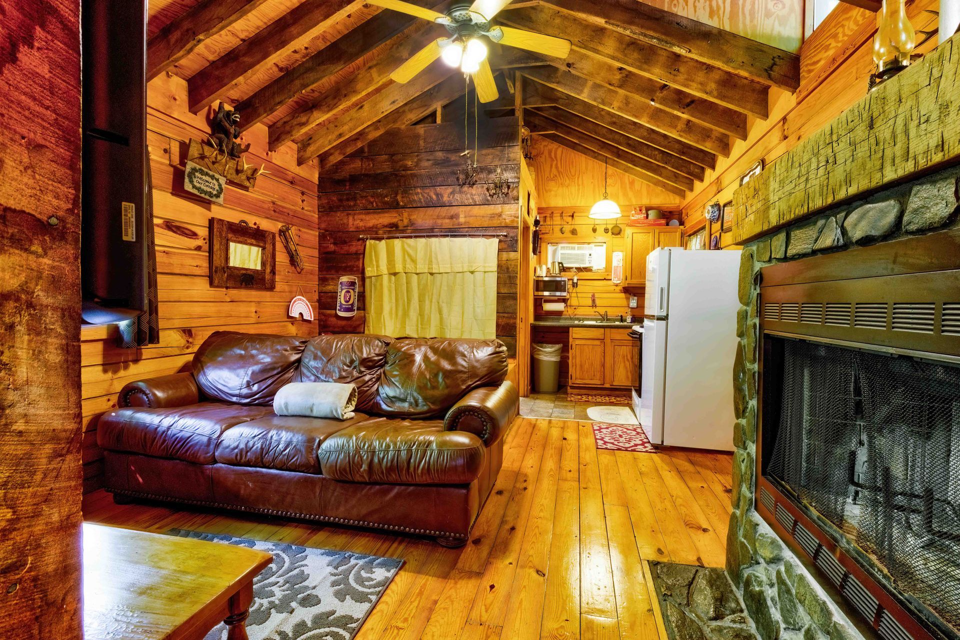 Rustic log cabin living room with leather sofa, wooden beams, fireplace, and kitchen area in the background