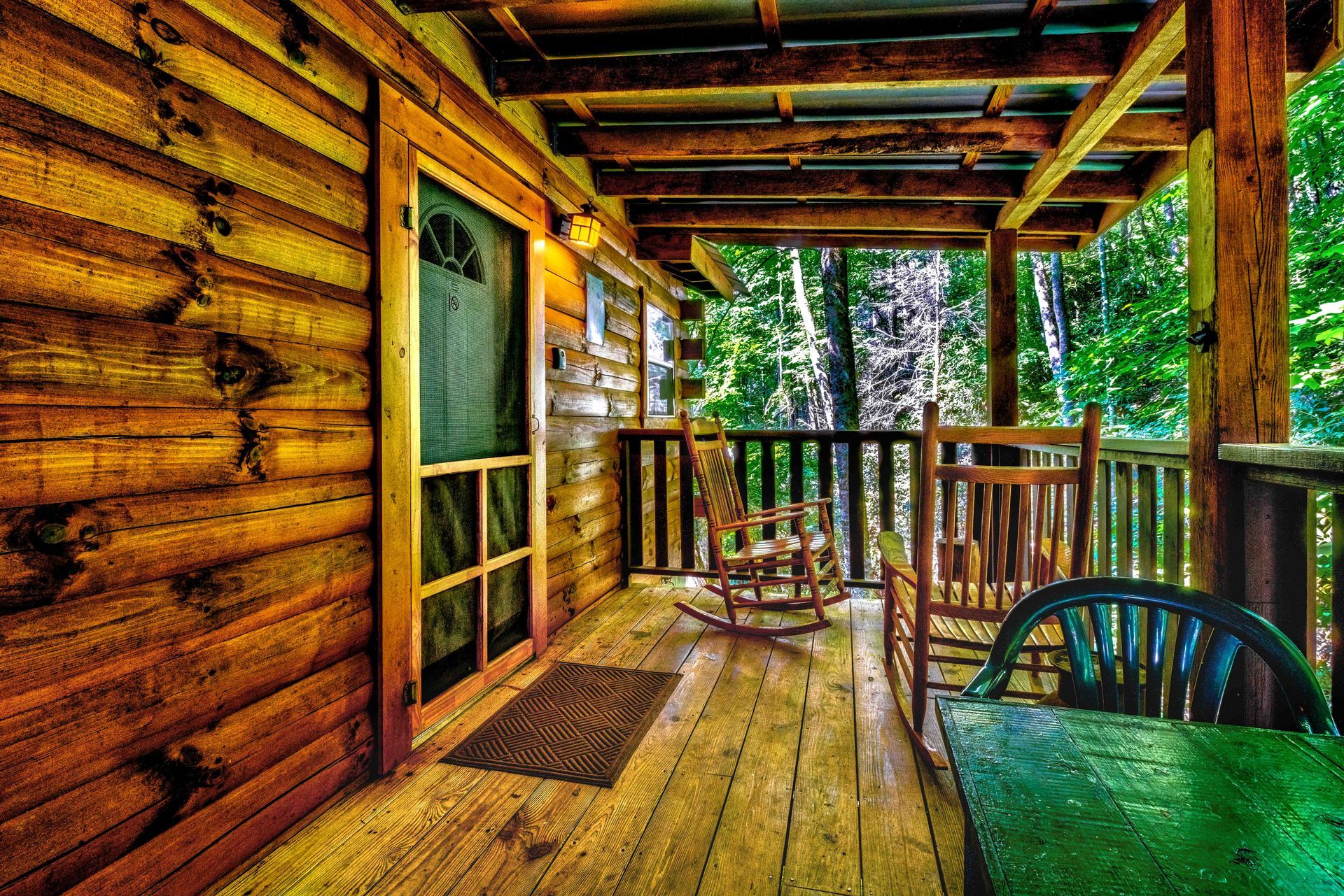 Wooden cabin porch with log walls, railing, and green table overlooking a wooded view
