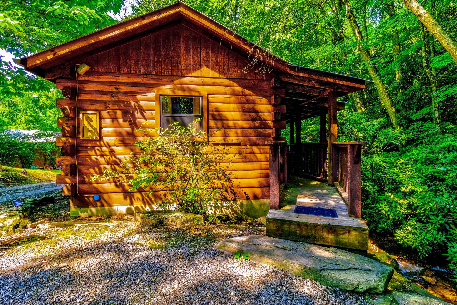 Small wooden cabin with a porch, surrounded by lush green trees and gravel in bright sunlight.