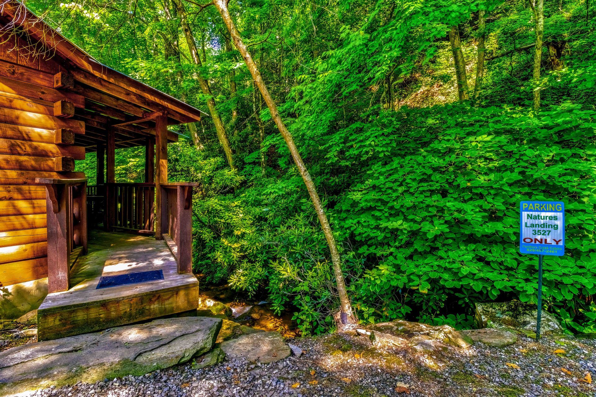 Wooden cabin porch by a forest trail, surrounded by lush green trees and a small sign nearby.
