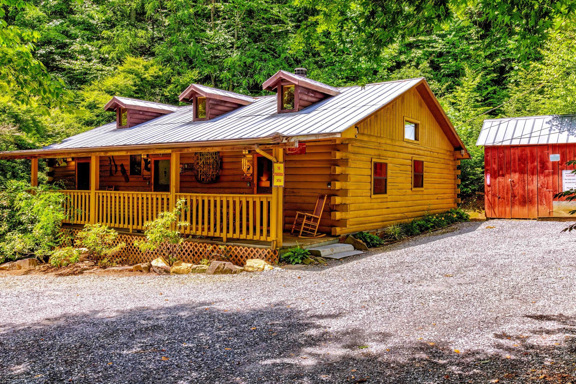 Rustic log cabin with porch and red shed beside a gravel driveway in a green wooded setting
