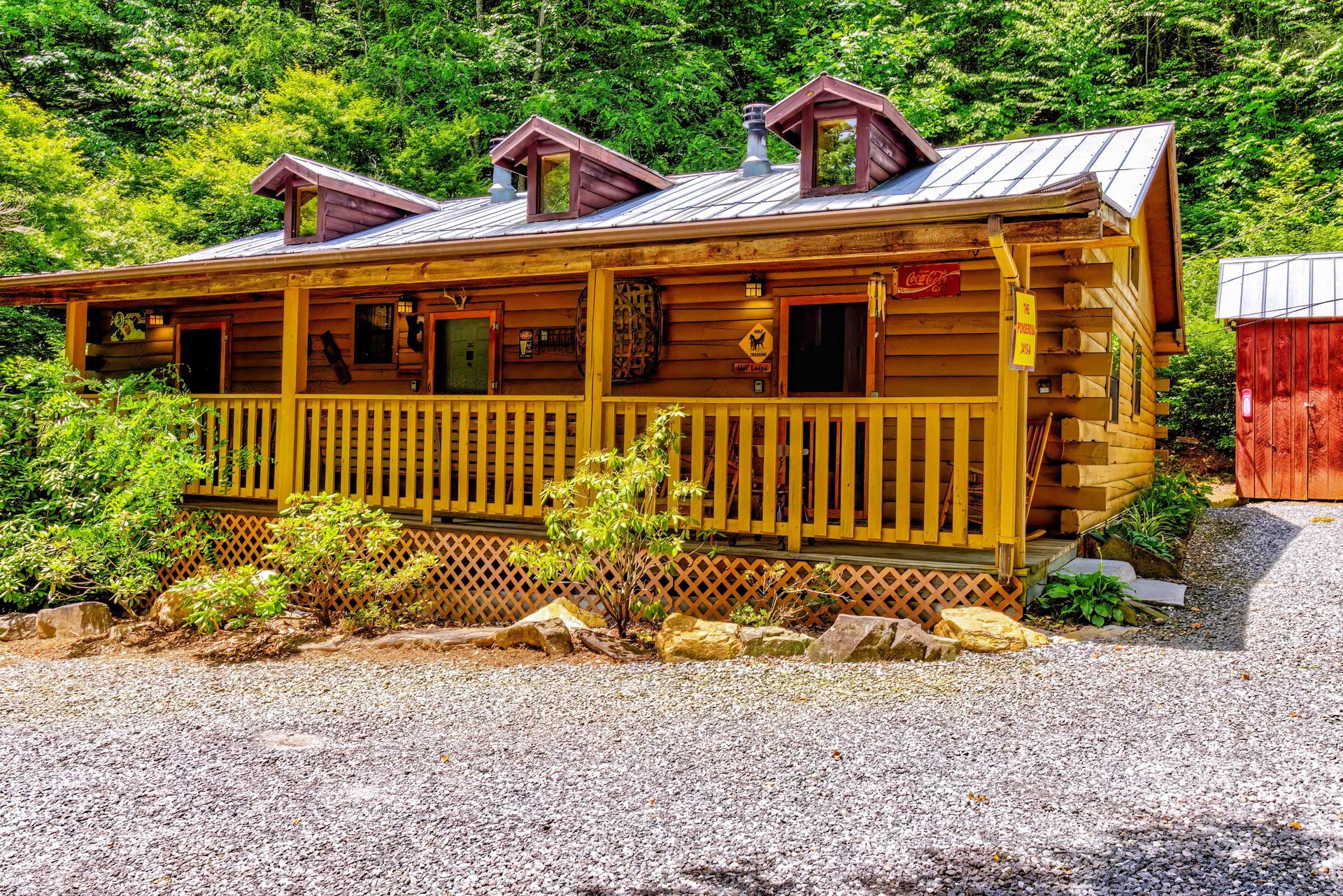 Rustic log cabin with porch and dormer windows surrounded by greenery and gravel driveway