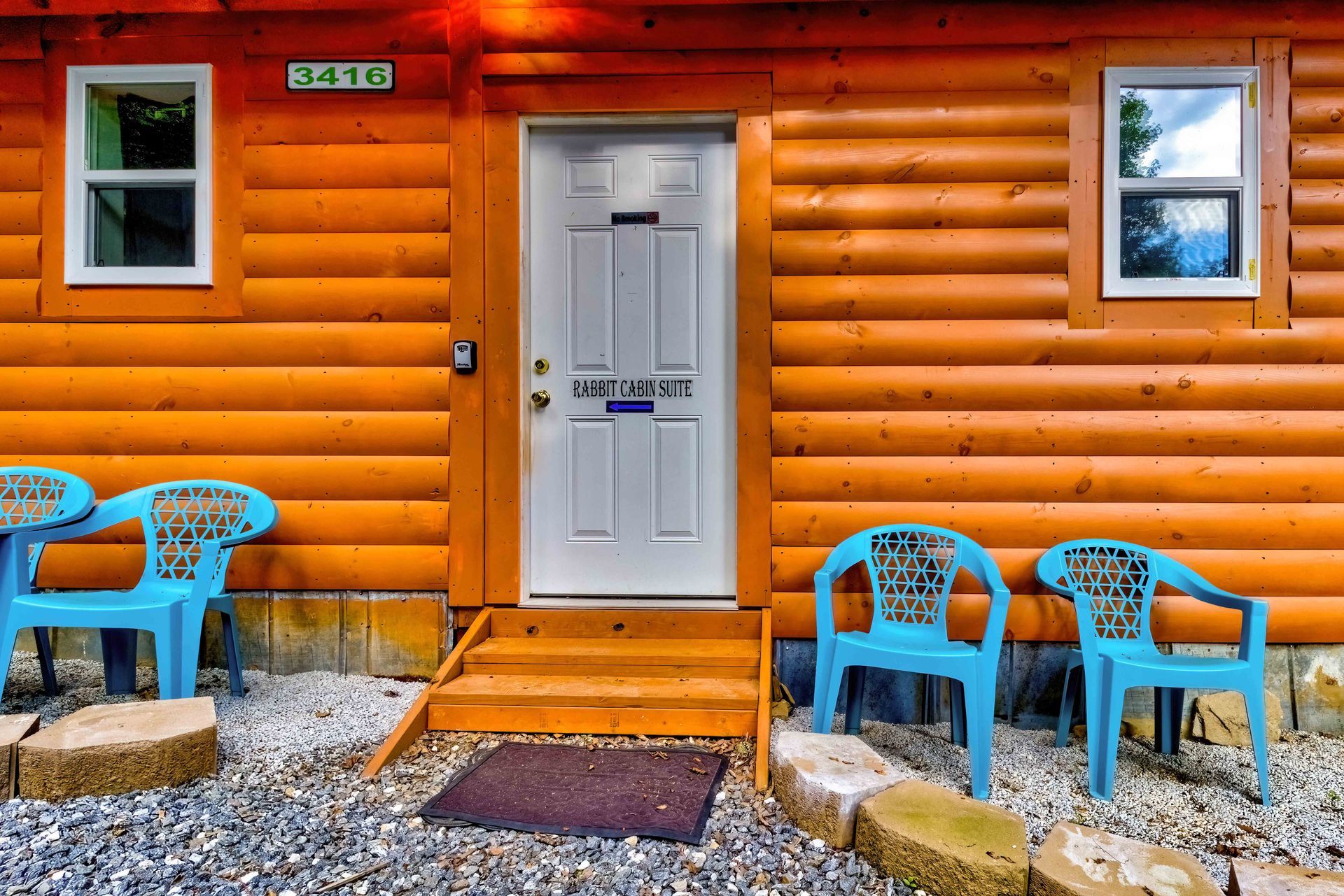 Wooden cabin entrance with white door, two windows, and blue chairs on a gravel porch