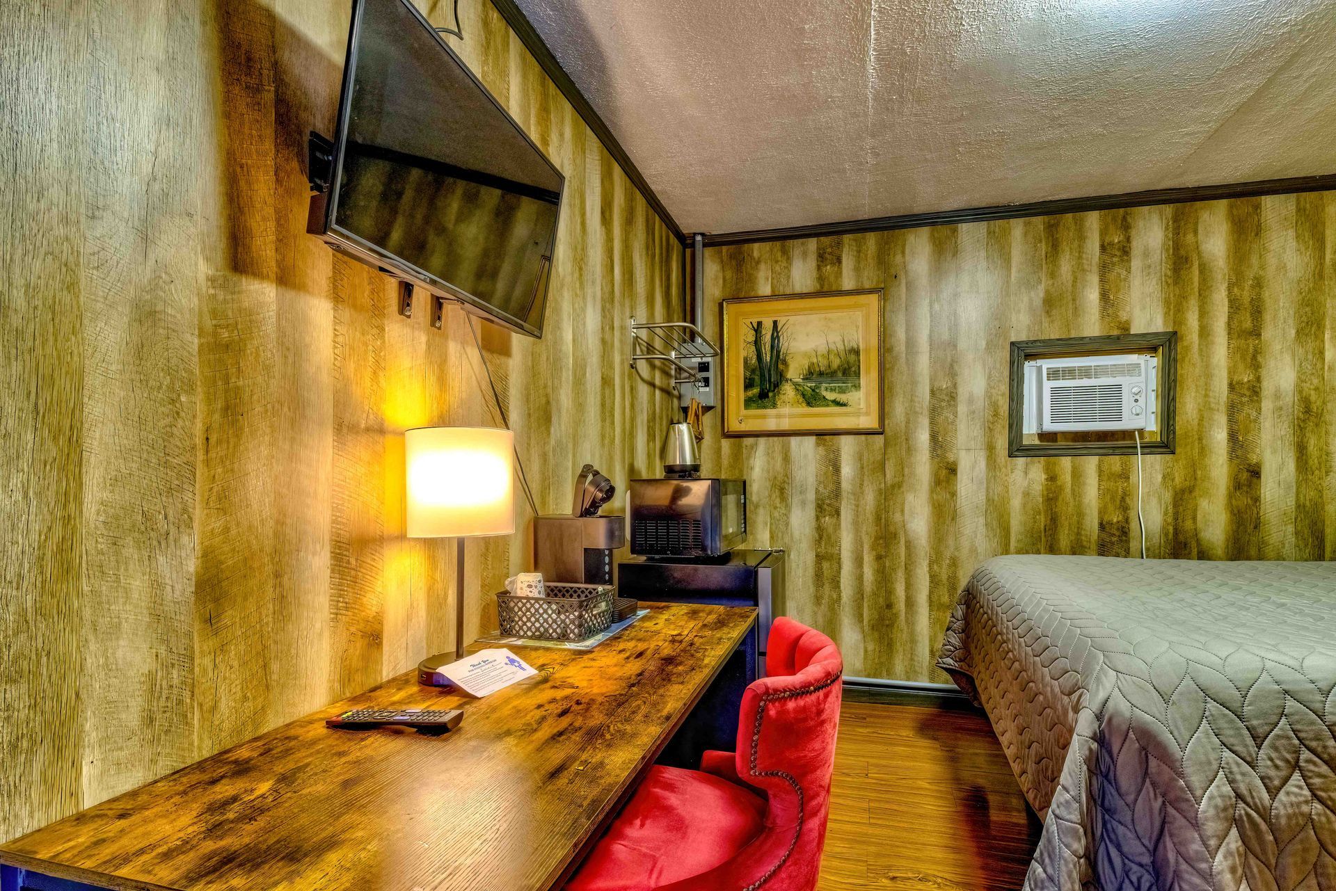 Small hotel room with a wooden desk, red chair, lit lamp, and bed against striped walls.