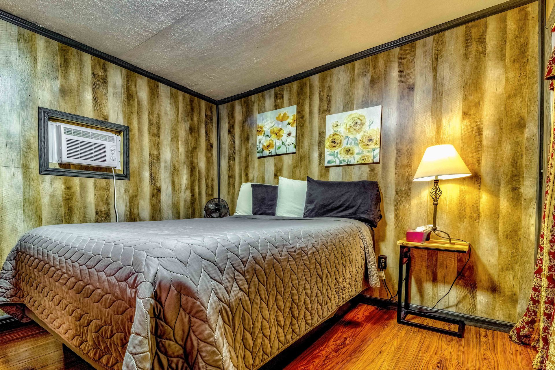 Cozy bedroom with a queen bed, wood-paneled walls, framed art, and a lit bedside lamp.