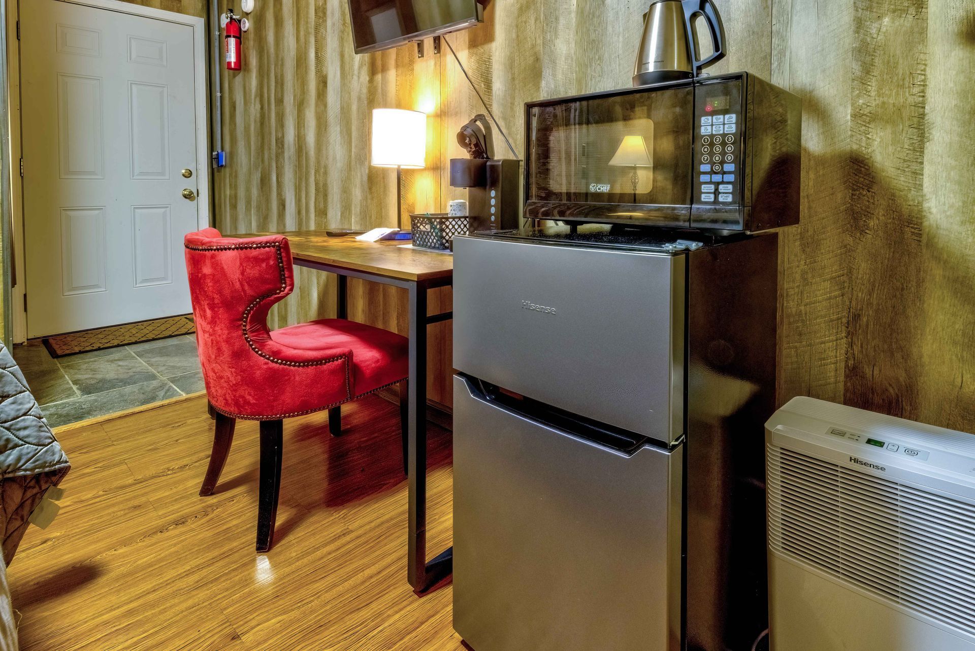 Small room with a mini fridge, desk, red chair, lamp, and microwave on a wooden floor
