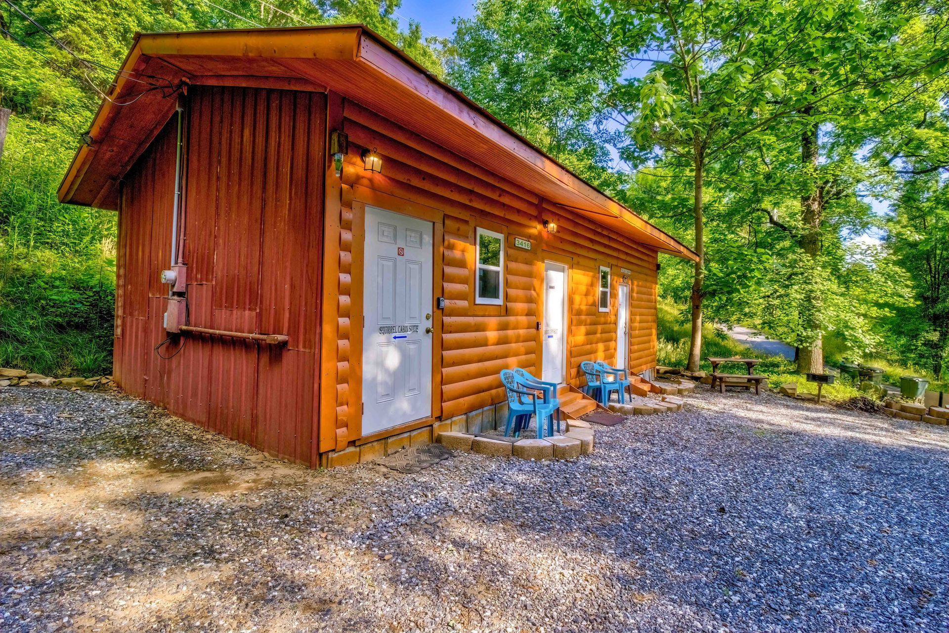 Small wooden cabin with a white door and blue chairs on a gravel lot, surrounded by trees.
