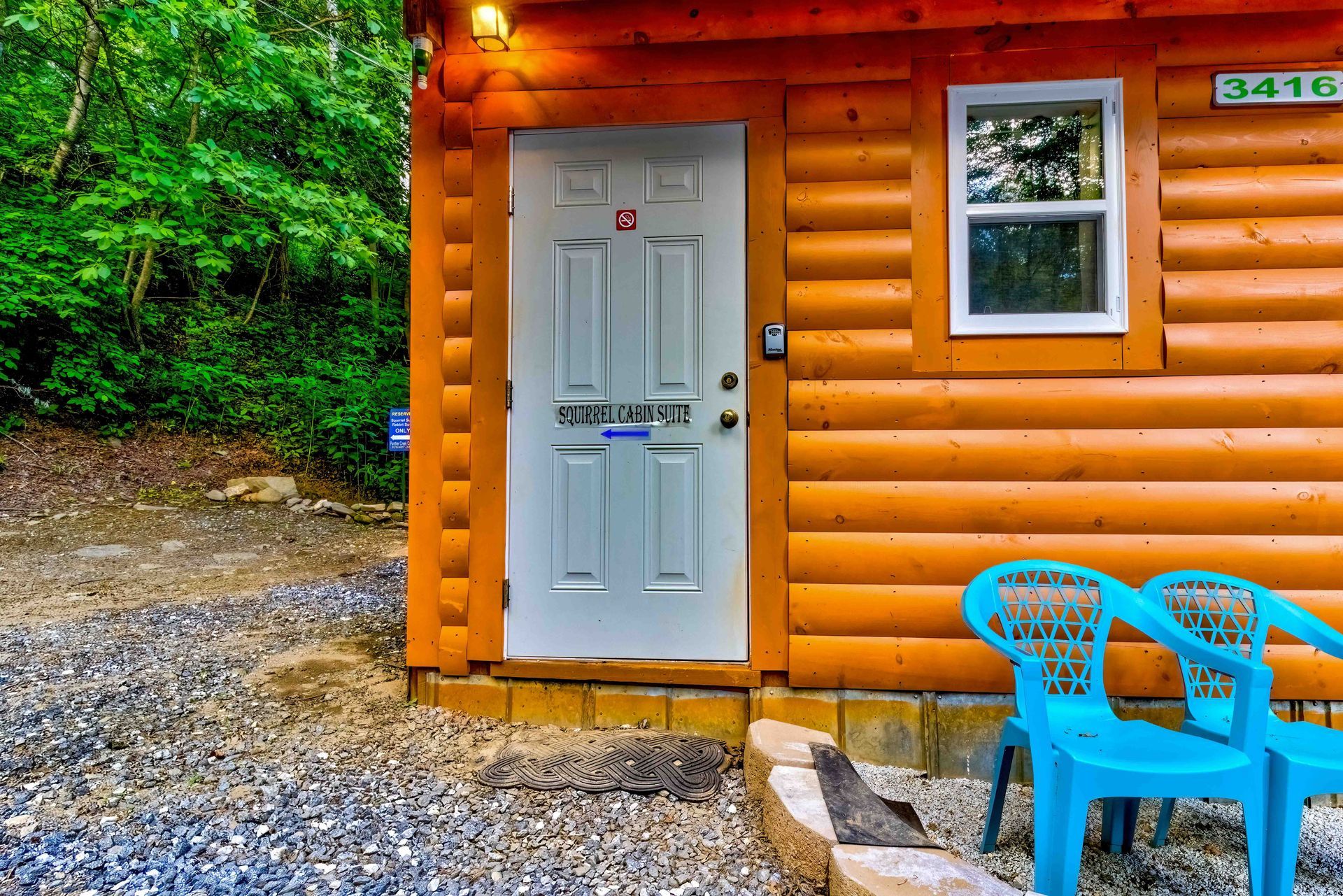 White cabin door on a log cabin with blue chairs on a gravel porch, surrounded by trees.