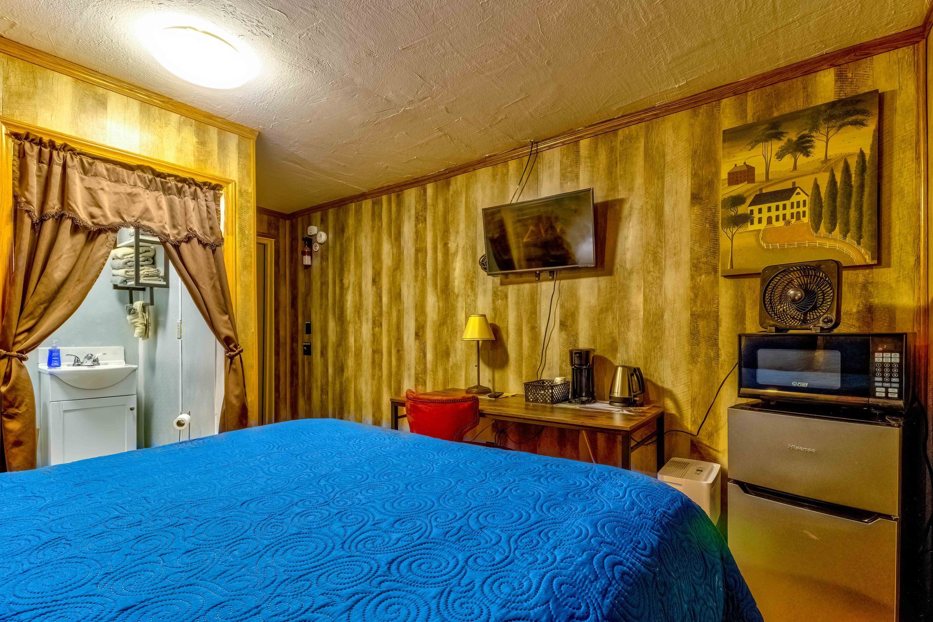 Small hotel room with blue bedspread, window curtains, dresser, TV, and mini fridge.