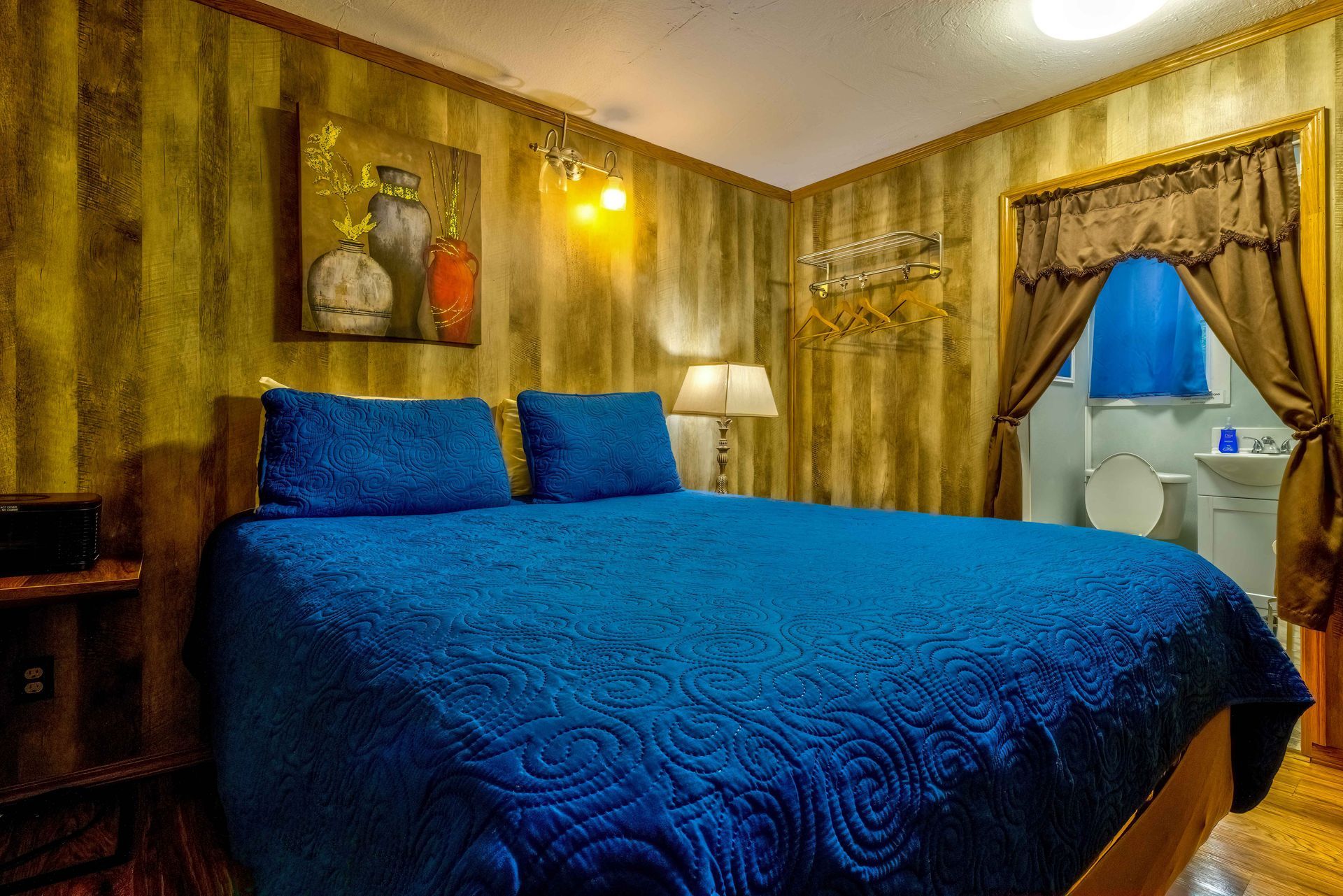 Blue bed in a cozy wood-paneled bedroom with lamps, framed art, and a window with curtains.