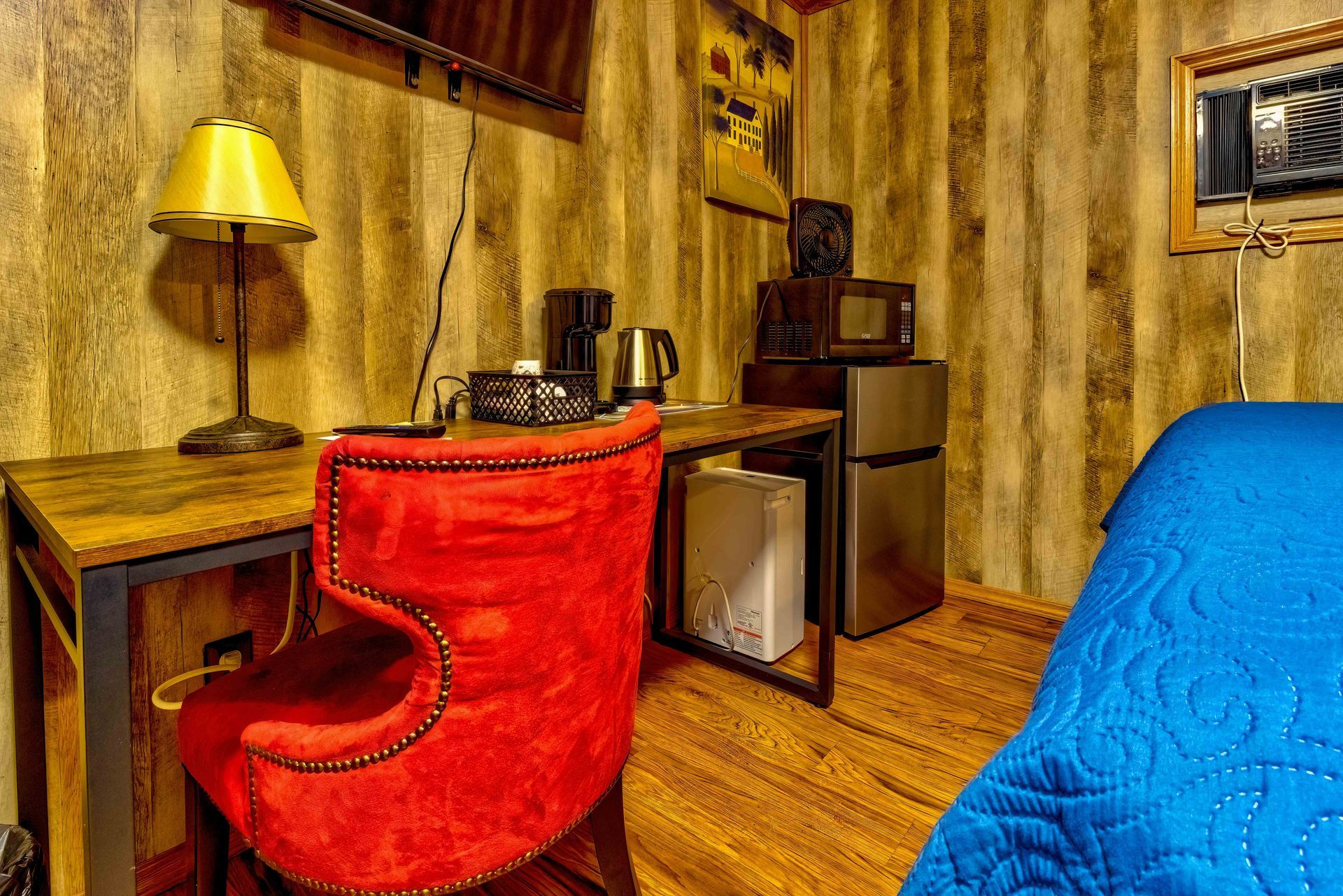 Cozy room with red chair, wooden desk, yellow lamp, mini fridge, and blue bedspread.