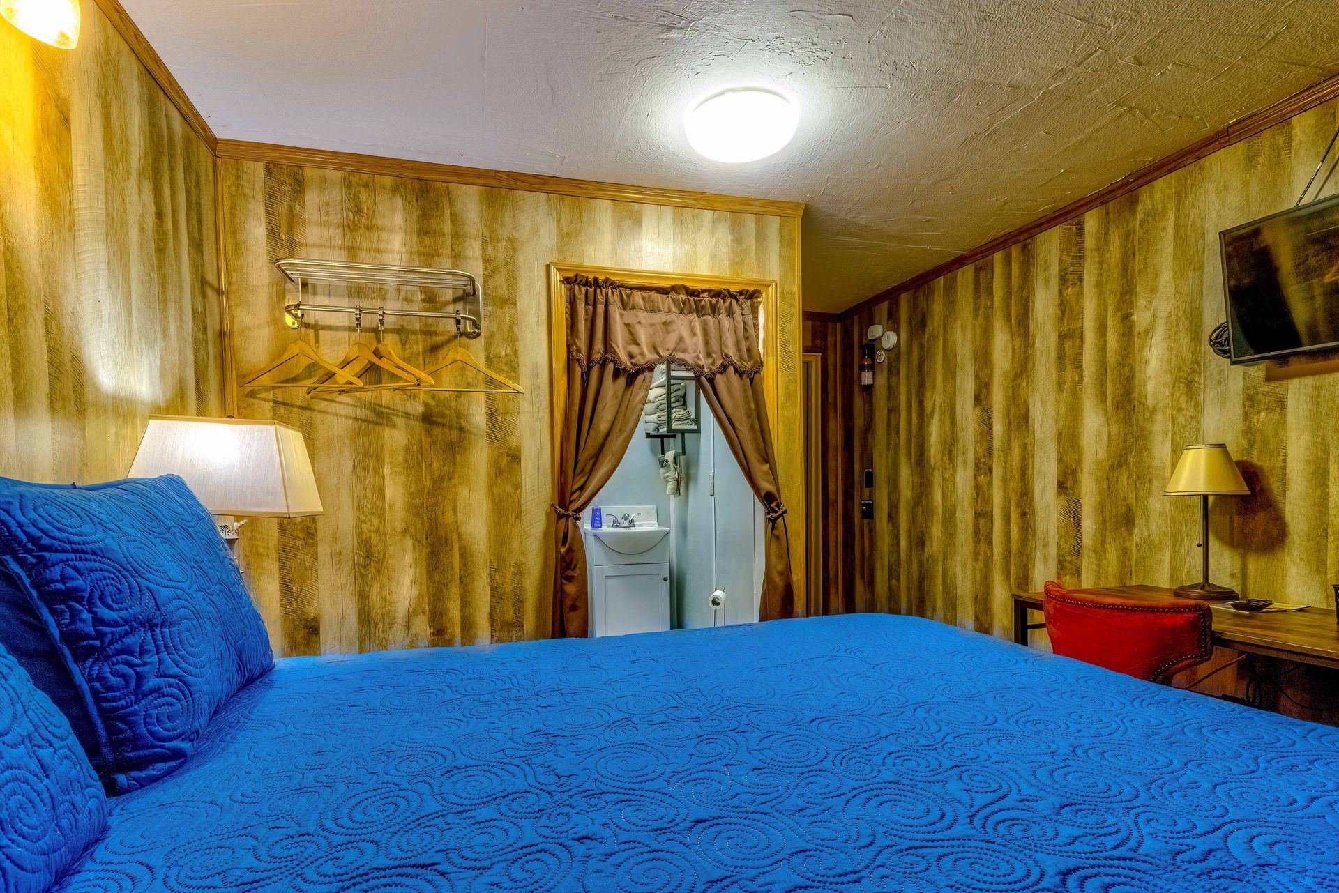 Blue bed in a yellow-walled bedroom with curtains, a lamp, and a wall-mounted TV.