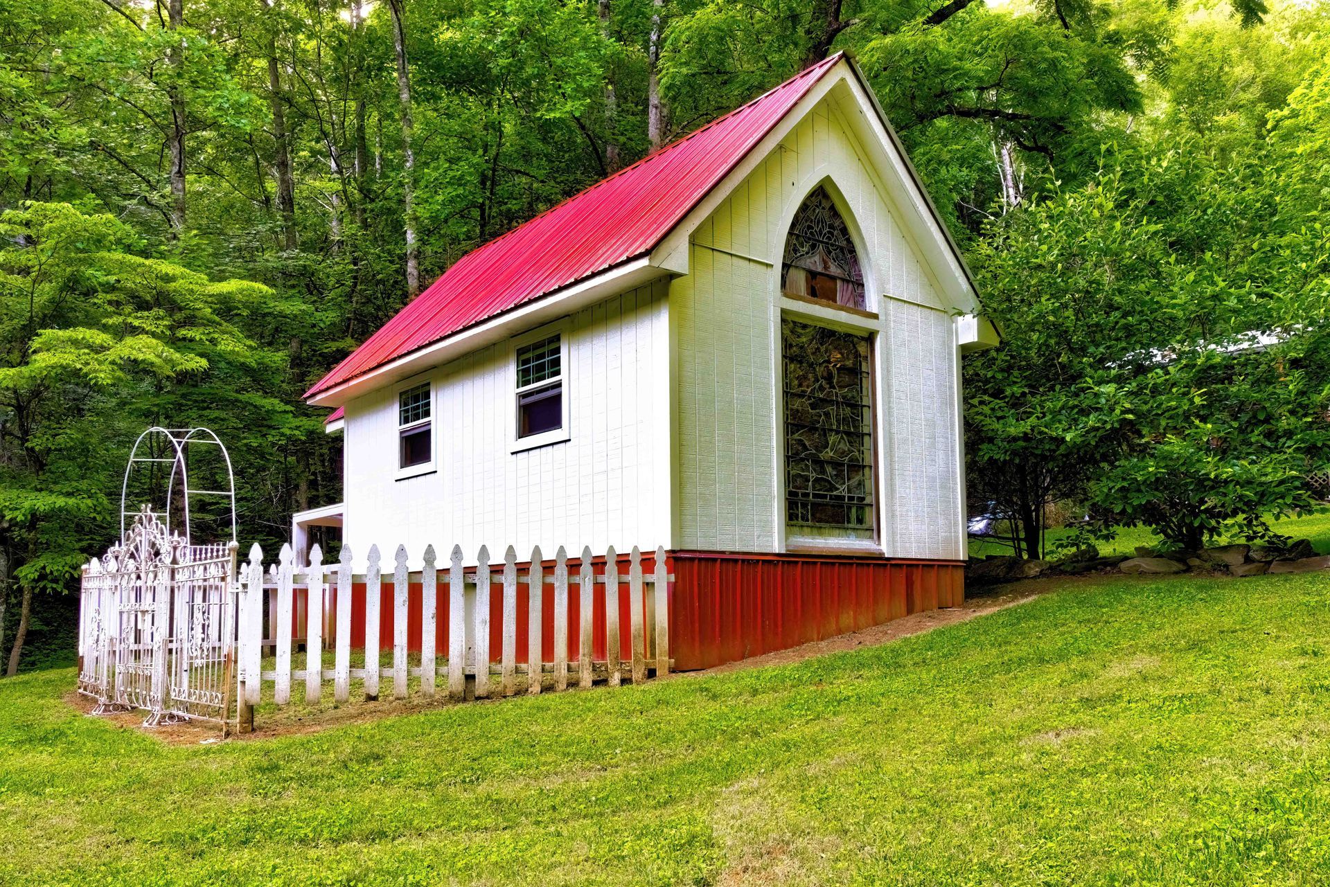 Small white chapel with a red roof and picket fence in a grassy, wooded setting