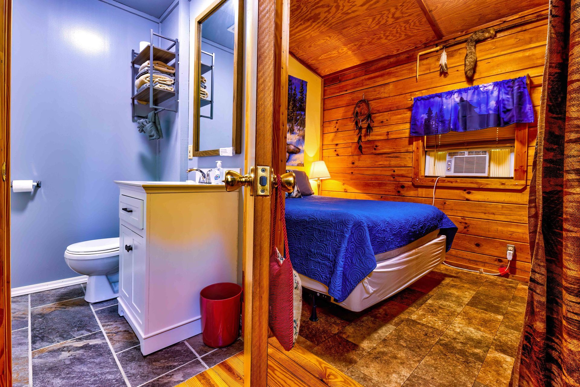 Small rustic bedroom with a bed, blue blanket, bathroom sink, and wooden walls under warm lighting