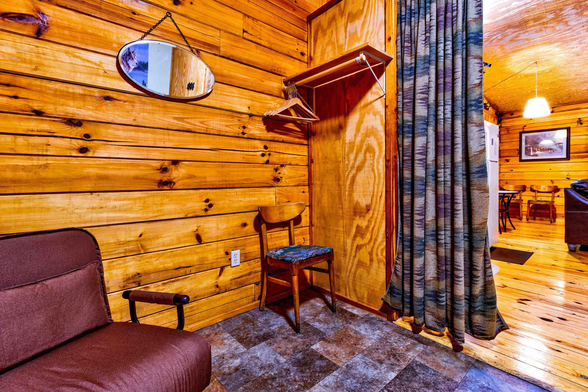 Cozy log cabin room with brown sofa, round mirror, small table, and striped curtains opening to a kitchen area