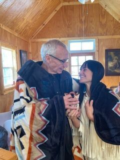 Two people smiling and holding hands inside a wooden cabin, wrapped in patterned blankets.