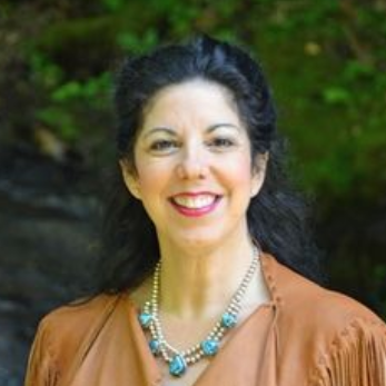 Smiling woman wearing a turquoise necklace and brown top, standing outdoors against a green background