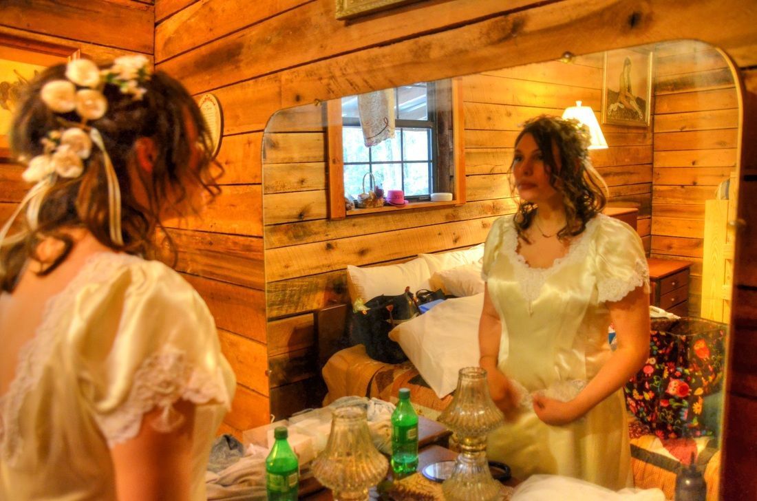 Woman in a white dress looking into a mirror in a cozy wooden room with flowers and a window.