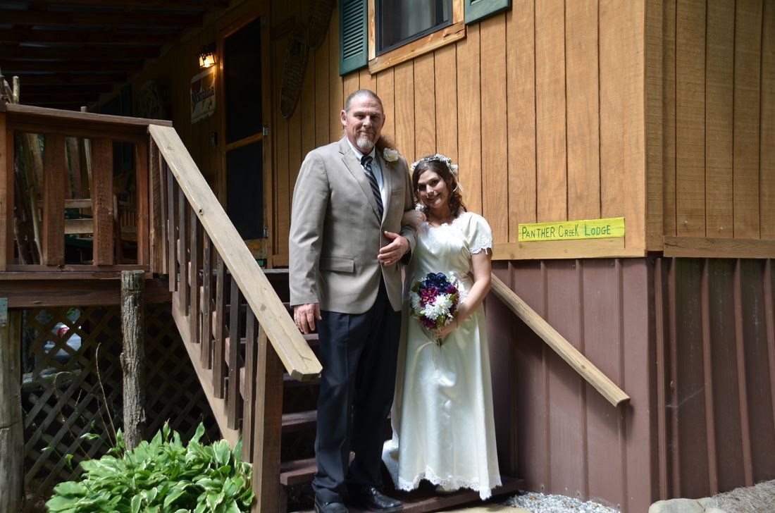 Bride and groom standing on wooden steps beside a rustic building, holding a bouquet.