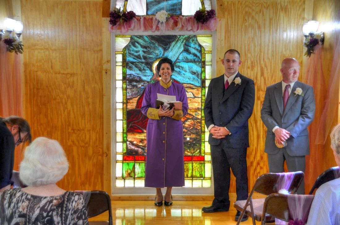 Woman at a lectern in a purple suit, with two men standing beside her on a small stage before stained glass.
