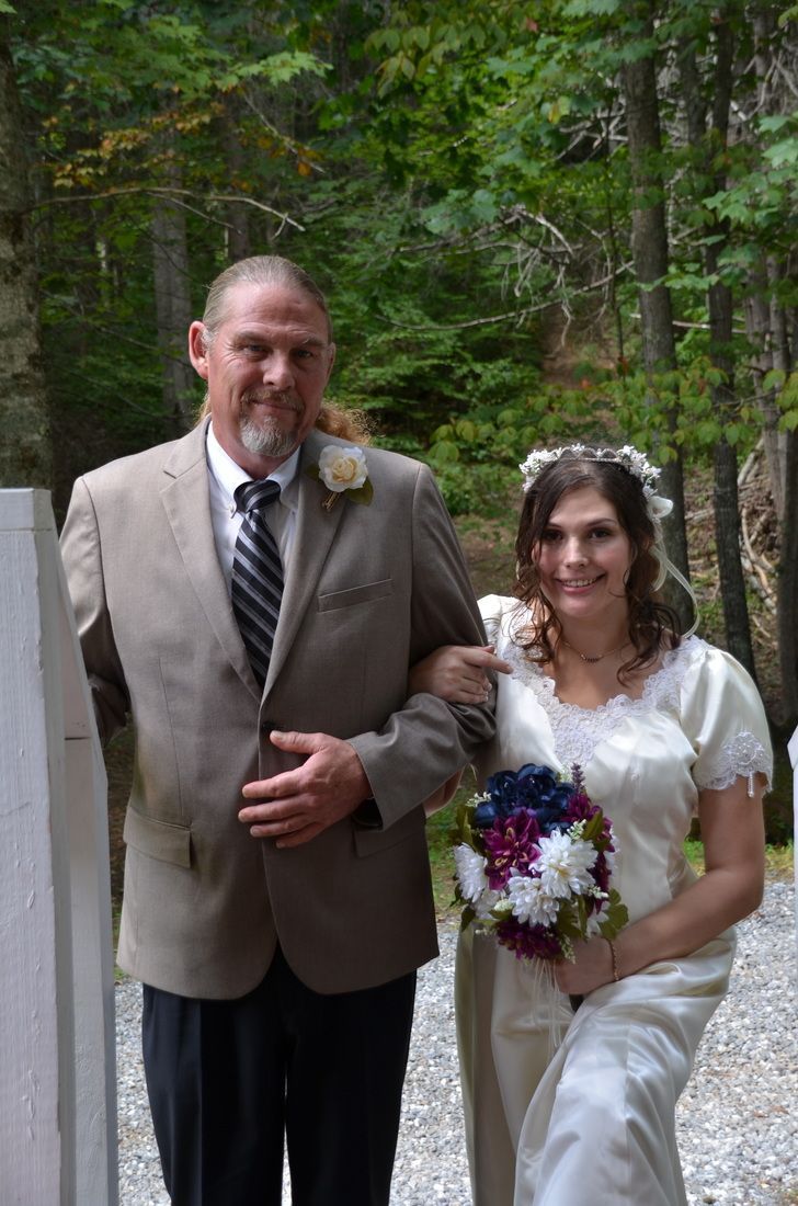 Bride in white dress with bouquet walks beside groom in tan suit on a wooded path