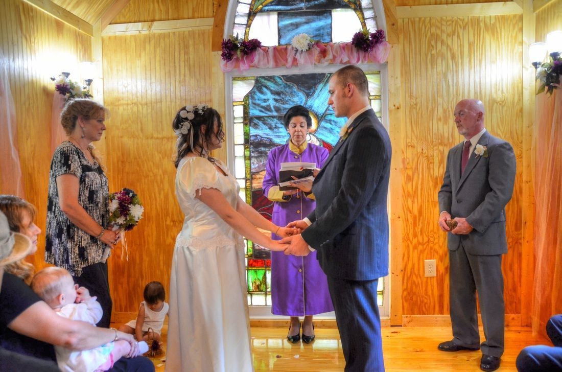 Wedding couple exchanging vows under a wooden canopy, with officiant and guests nearby.
