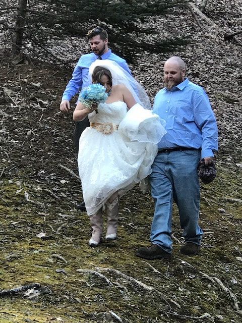 Three adults walking on a rocky path, one carrying a bride in a white dress holding a blue bouquet.