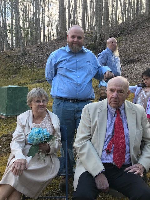 Three people posed outdoors in a wooded setting, with one holding a bouquet and two seated in formal clothes.