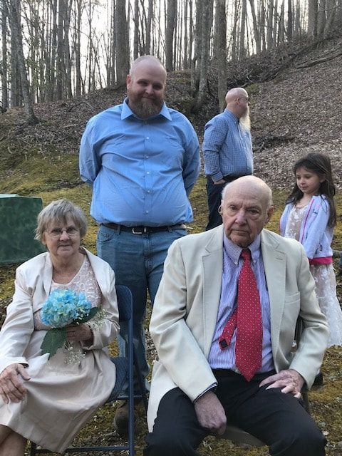 Group seated and standing outdoors on a wooded hillside, including a bouquet and formal clothing