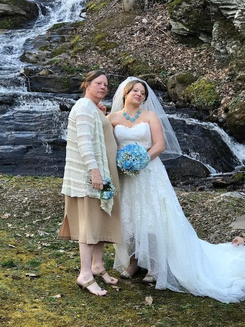 Two people in wedding attire standing in front of a waterfall, one holding a blue bouquet.