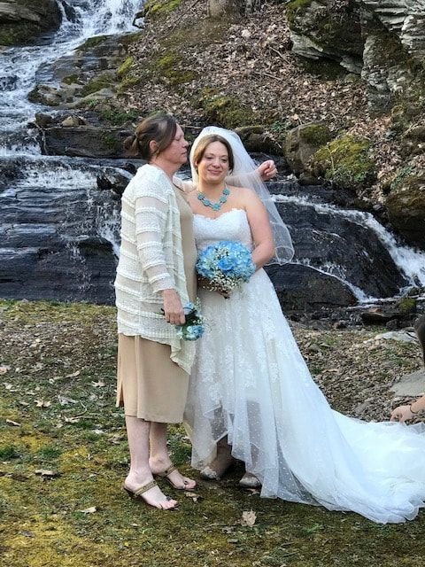 Two people in wedding attire smiling by a rocky waterfall, with a blue bouquet and long white veil