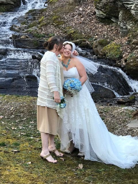 Bride and companion kissing beside a waterfall, with a long white gown and blue bouquet