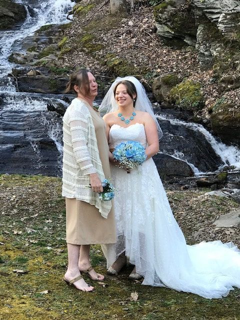 Two women in wedding attire standing together by a waterfall, smiling and holding bouquets.