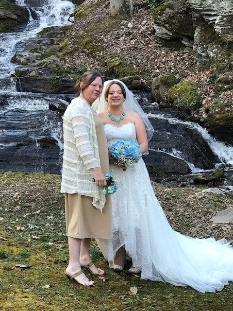 Two women in wedding attire posing by a rocky waterfall, one holding a blue bouquet