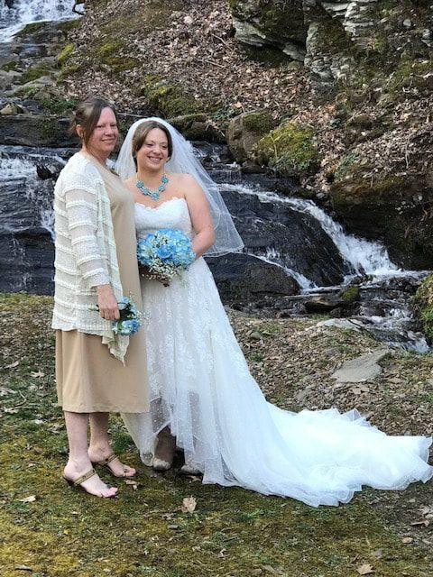 Two women in formal dresses stand smiling by a small waterfall outdoors.