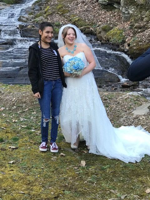 Bride in a white gown holding a blue bouquet stands beside a smiling person on a rocky hillside by a waterfall