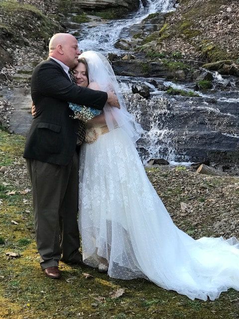 Bride and groom embracing beside a small waterfall, with the bride’s long white veil trailing on the grass.