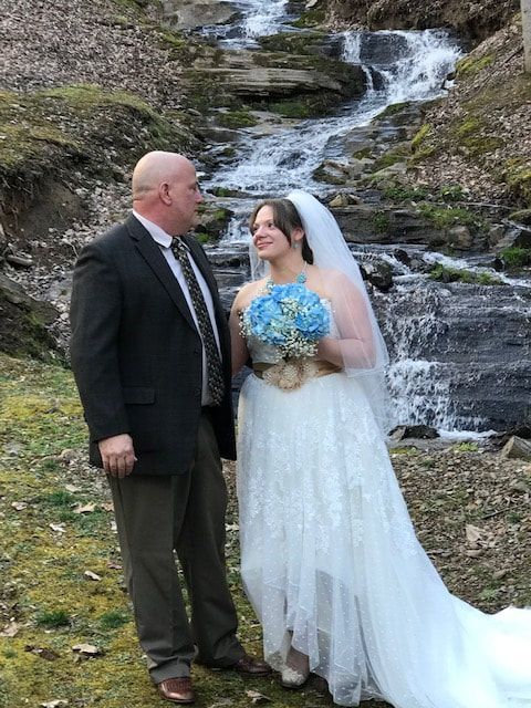 Bride and groom posing by a waterfall, bride holding a blue bouquet and smiling in a white gown and veil