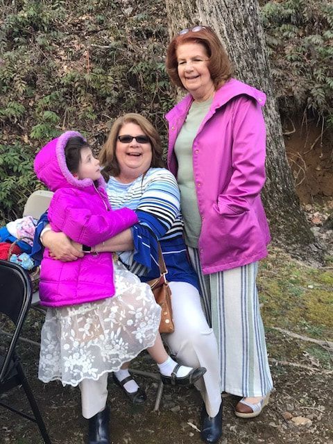 Three people outdoors, two in pink jackets, one seated in a wheelchair, smiling beside a rocky hillside