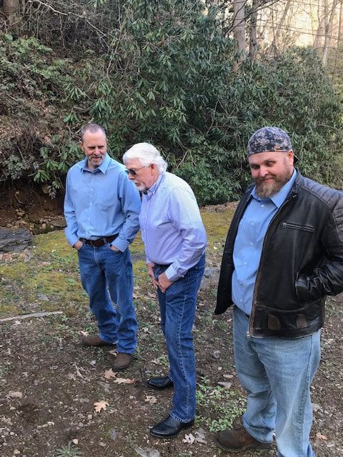 Three men standing outdoors on a dirt path beside dense bushes and trees, one bending forward.
