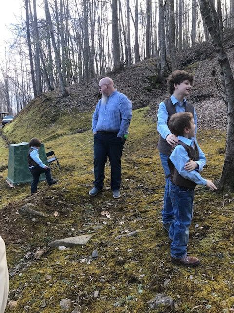 People standing and sitting on a wooded hillside path, with a green trash bin nearby