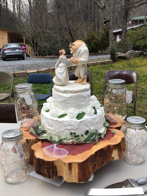 White tiered wedding cake topper on a tree-slice stand, surrounded by jars and chairs outdoors