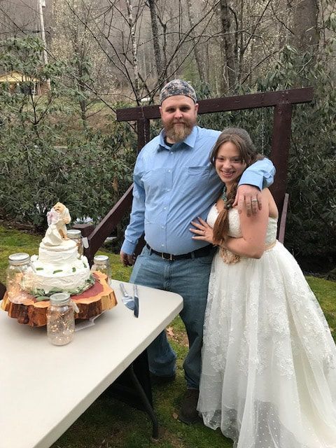 Two people posing beside a cake table at an outdoor celebration, with trees in the background.