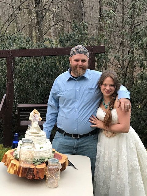 Man and young woman pose beside a decorated cake outdoors at a wooded celebration.