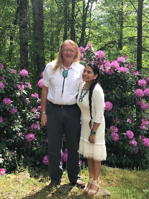 Two people standing in front of pink flowering bushes in a wooded garden area.