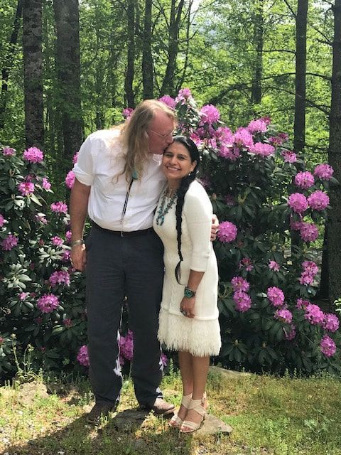 Two people standing together in a forest, surrounded by pink flowers, smiling and leaning close.