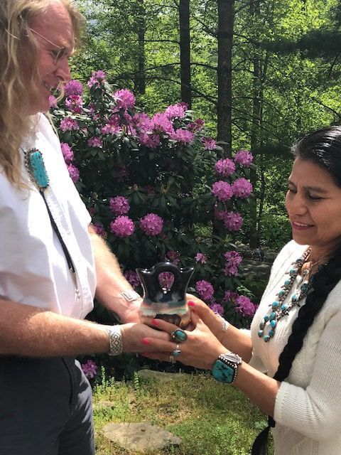 Two people exchange a small gift outdoors, with pink flowering bushes in the background.