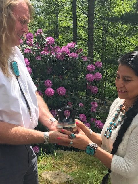 Two women exchanging a small black object in a garden with pink flowers.