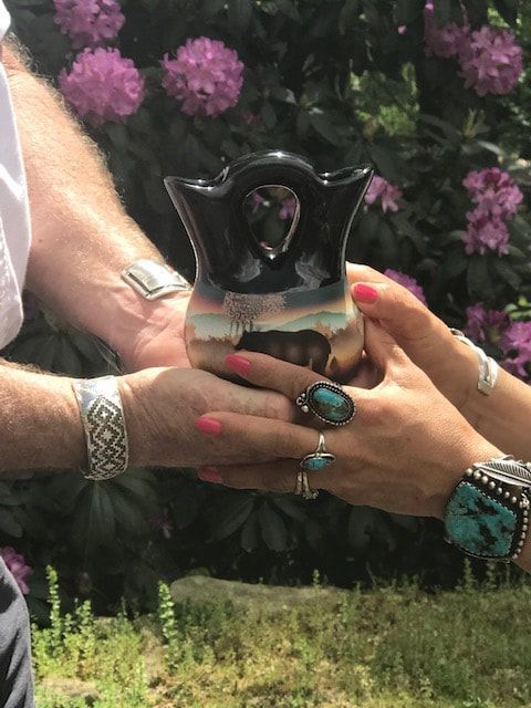 Hands holding a small black decorative object with an owl shape, outdoors with pink flowers in the background